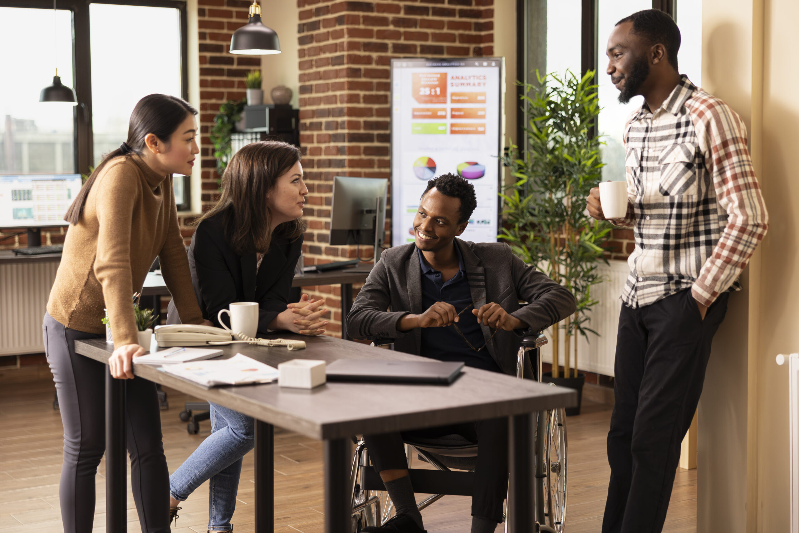 A diverse group of four colleagues smiling and chatting during a break in a modern, brick-walled office, including a man using a wheelchair.